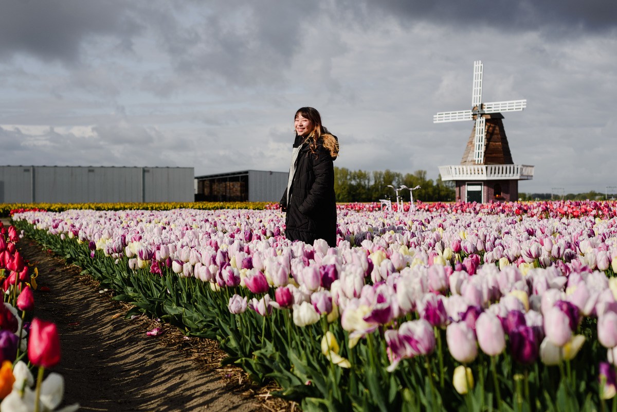 Portrait of a woman smiling among rows of tulips in Hillegom, wearing a black coat, with a windmill visible behind her after a stormy sky clears.