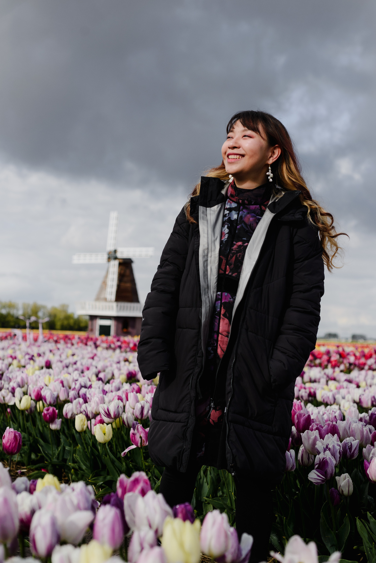Close-up portrait of a woman in a tulip garden, smiling softly with a windmill blurred in the background and dramatic spring sky overhead.