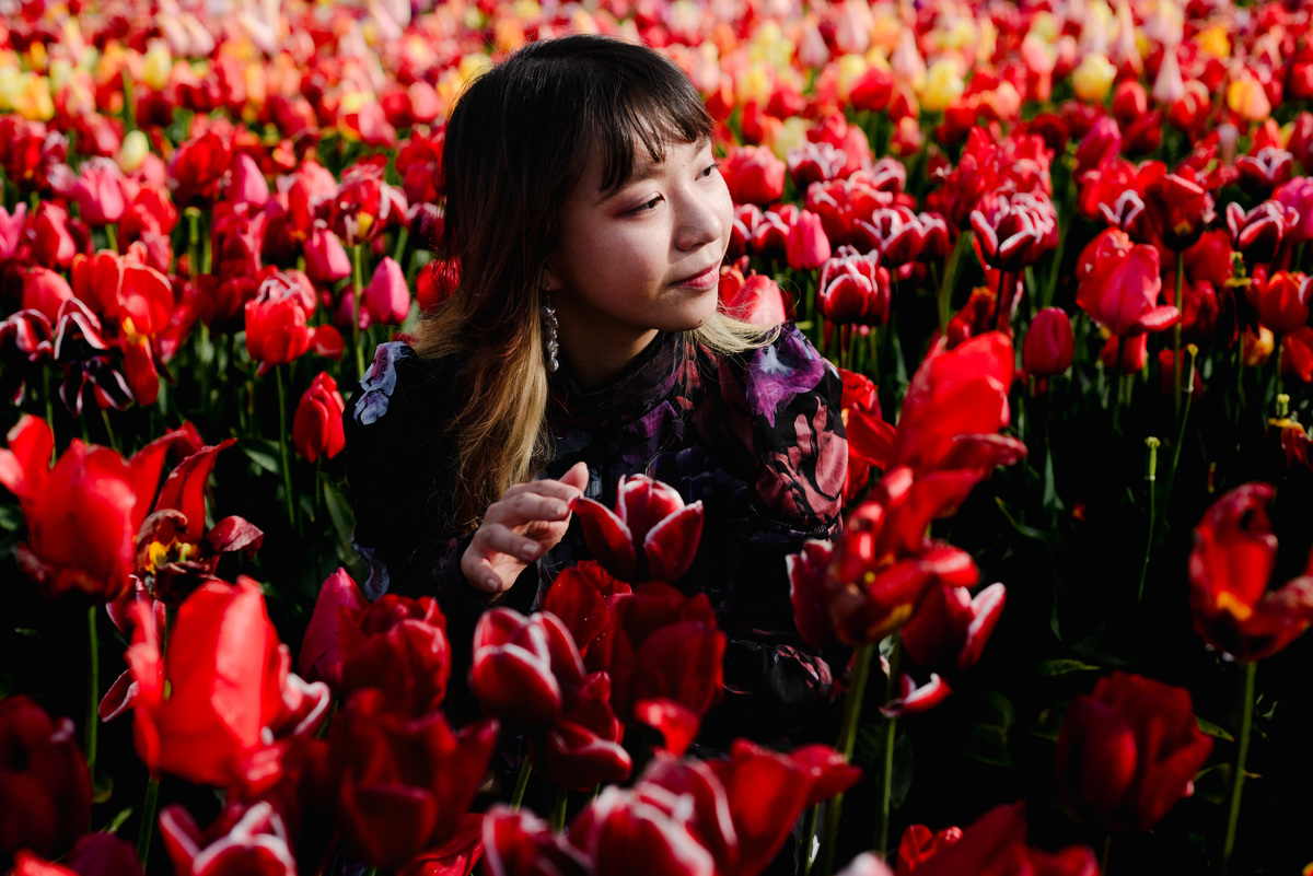 Close portrait of a woman surrounded by bright red tulips, looking to the side, with deep colors enhanced by post-rain light.