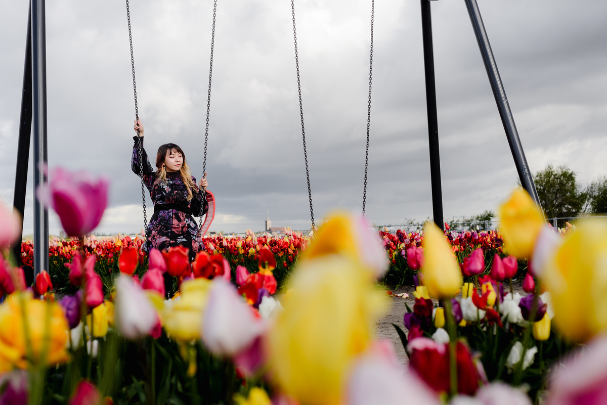 Wide shot of a woman on a swing surrounded by yellow, red, and pink tulips, with an open sky and quiet garden atmosphere.
