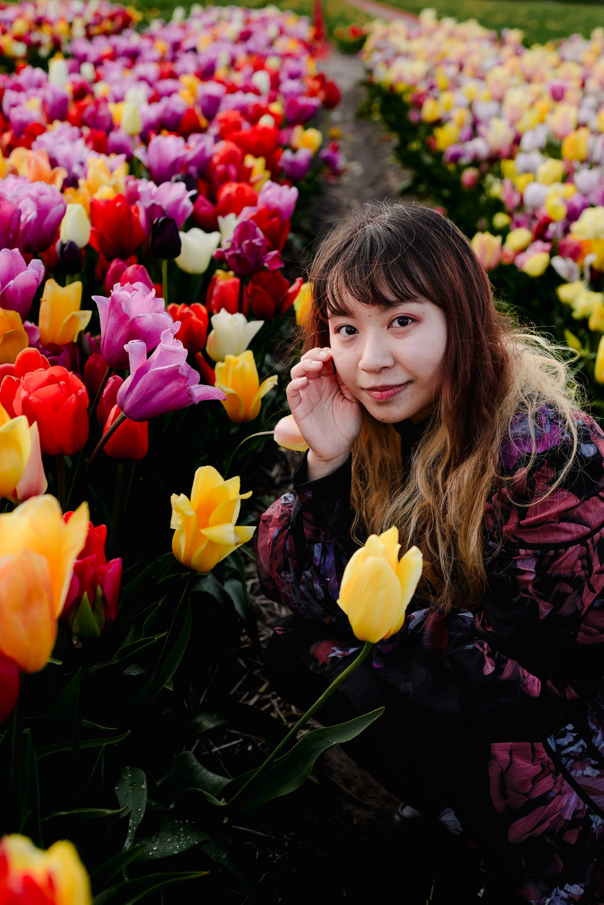 Close-up portrait of a woman crouching between tulip rows, surrounded by pink, red, and yellow tulips.