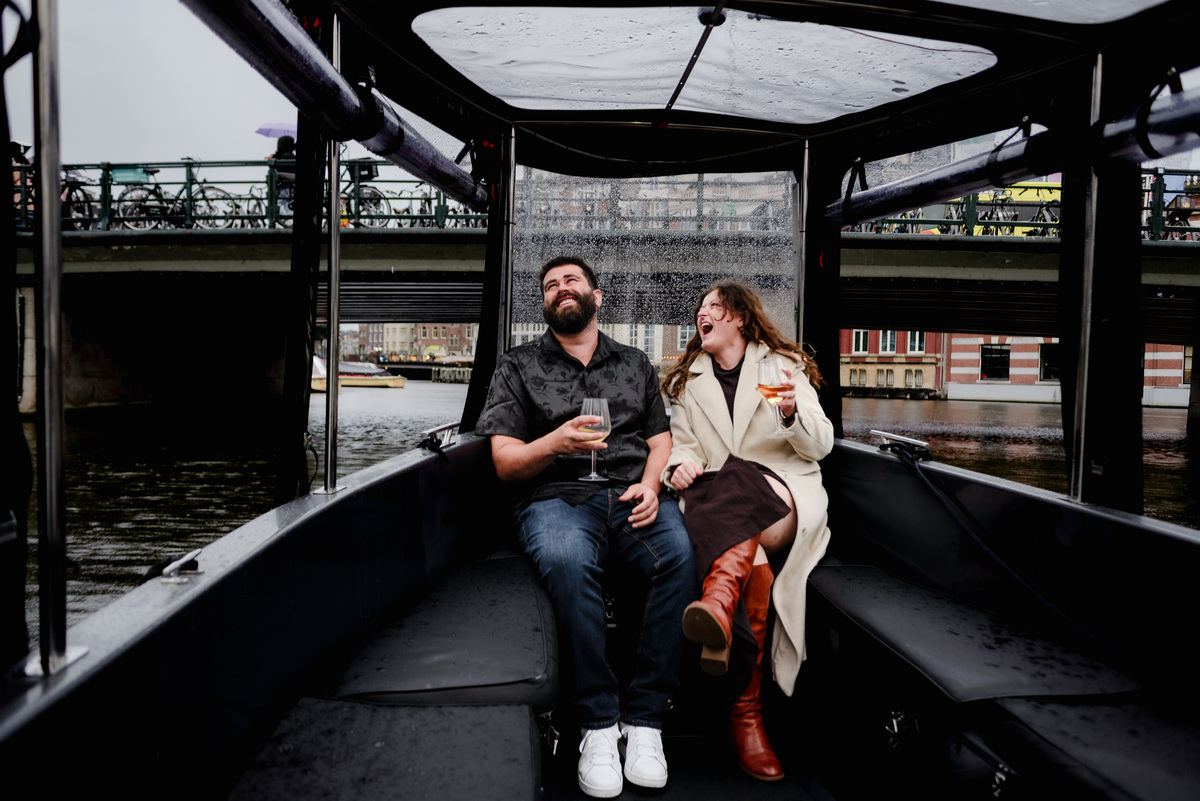 Engaged couple laughing and holding hands on the deck of a boat, surrounded by Amsterdam’s beautiful waterways.
