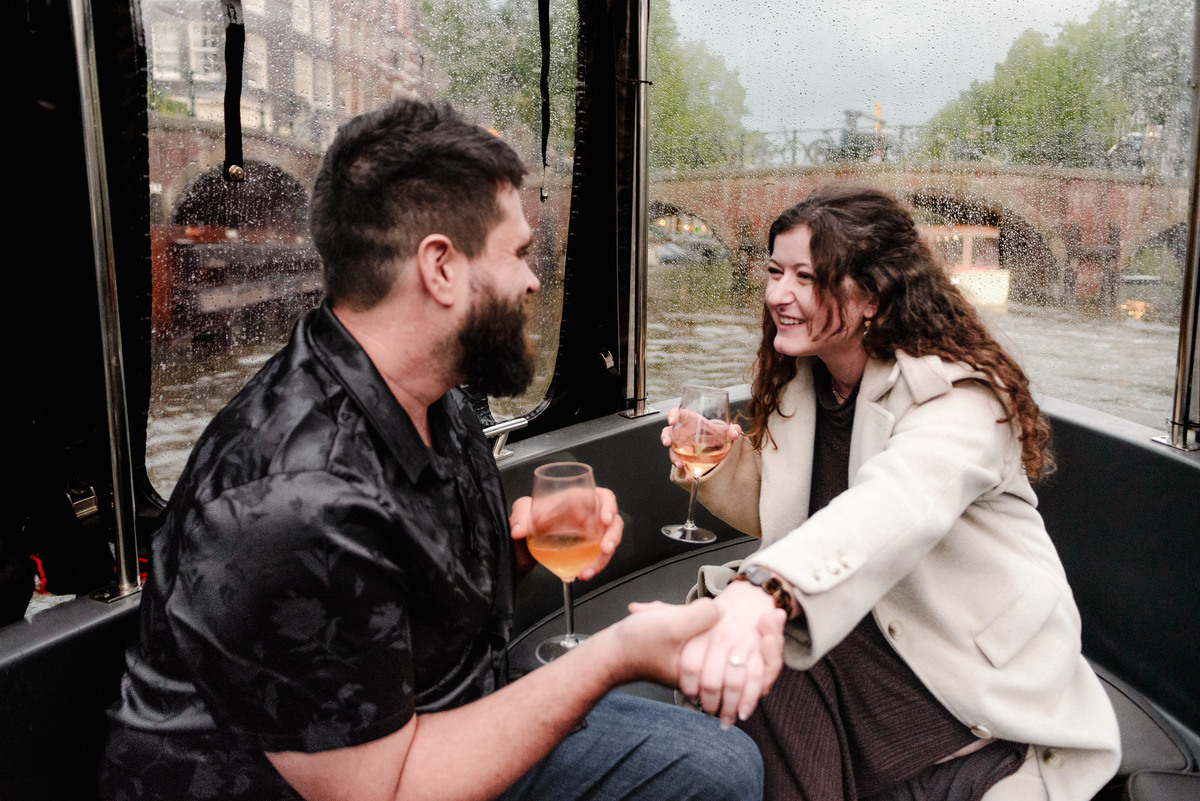Engaged couple laughing and holding hands on the deck of a boat, surrounded by Amsterdam’s beautiful waterways.