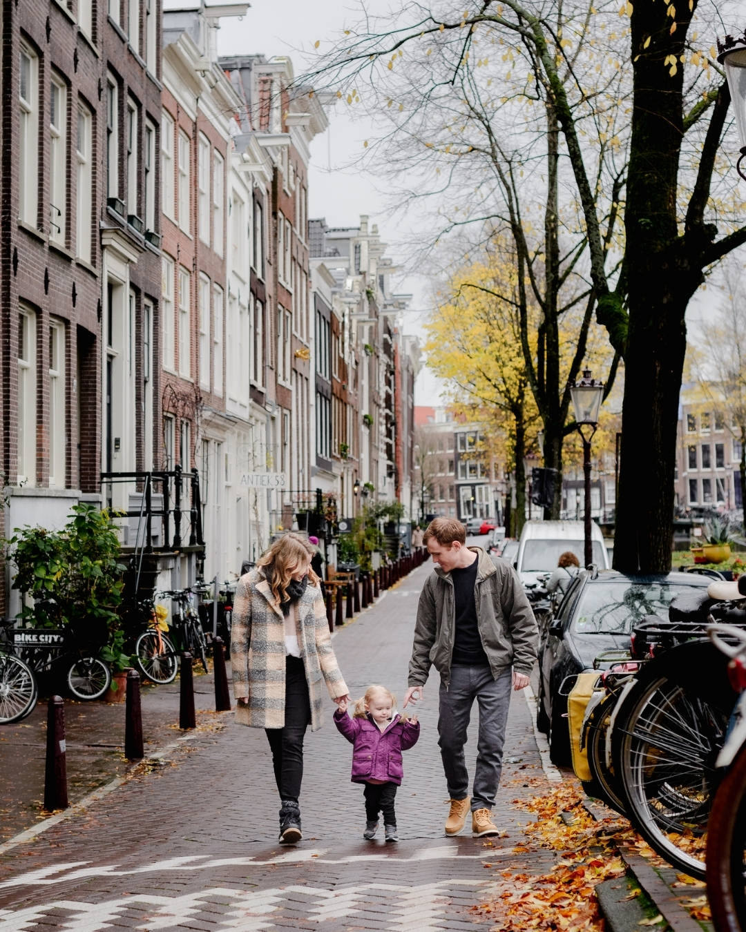 A family walks hand in hand through the charming streets of Jordaan, Amsterdam, during a cozy autumn photo session. With historic canal houses, golden leaves, and bicycles lining the streets,this moment captures the warmth of family adventures in the city