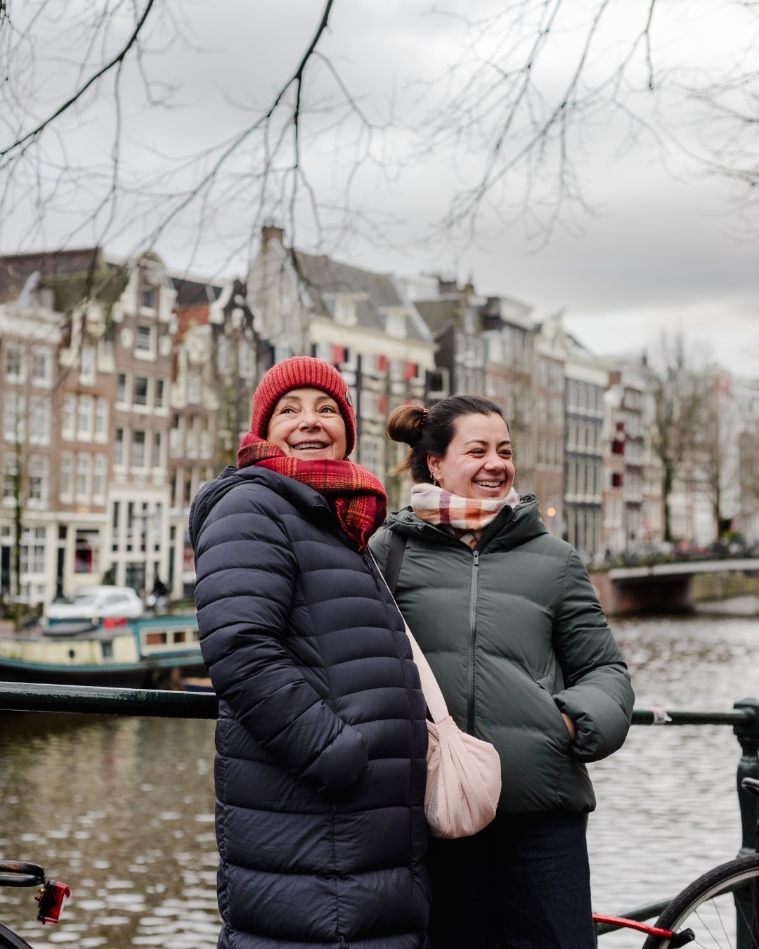 Standing by one of Amsterdam’s iconic canals, the two women share a joyful moment, wrapped in scarves and winter coats, with historic canal houses forming a picturesque background