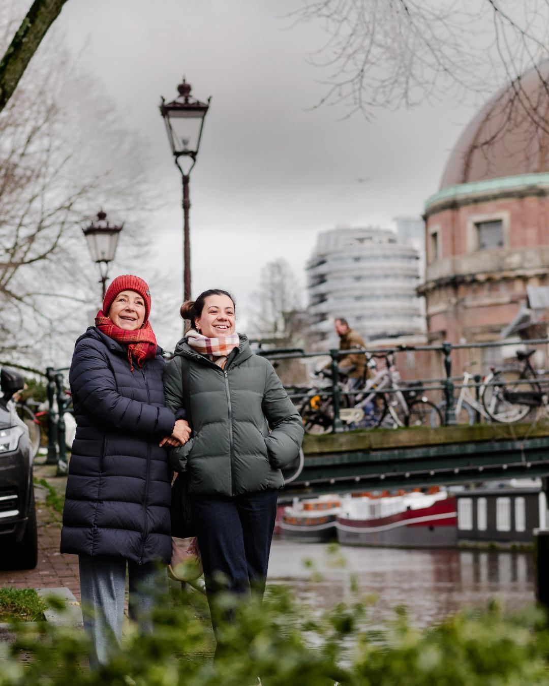 Embracing History and Modernity – In front of a historic Amsterdam landmark, the mother and daughter link arms, looking delighted by their surroundings. The old-world charm of the city comes alive in this candid moment.