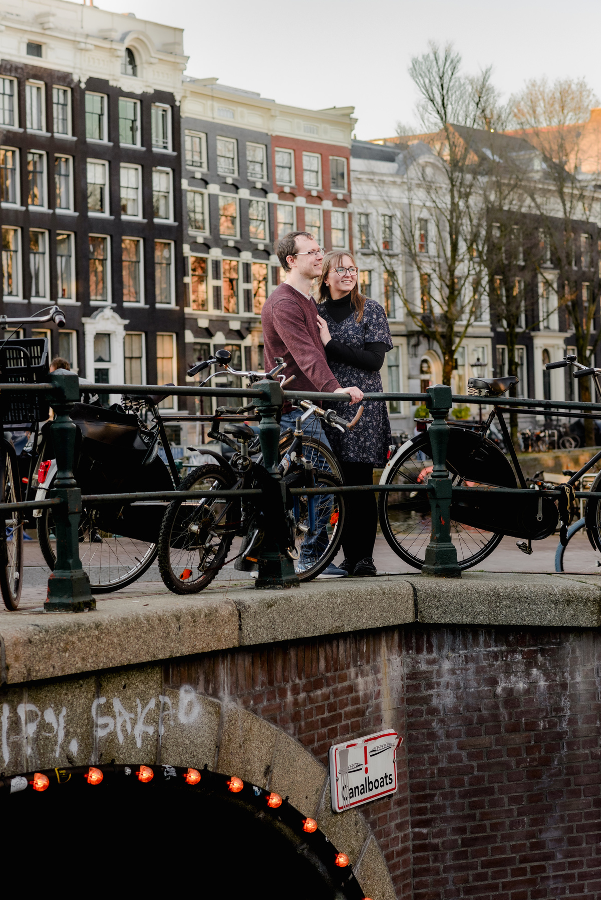 Couple enjoying a quiet moment on a bridge above an Amsterdam canal at sunset, surrounded by bicycles and soft golden light.