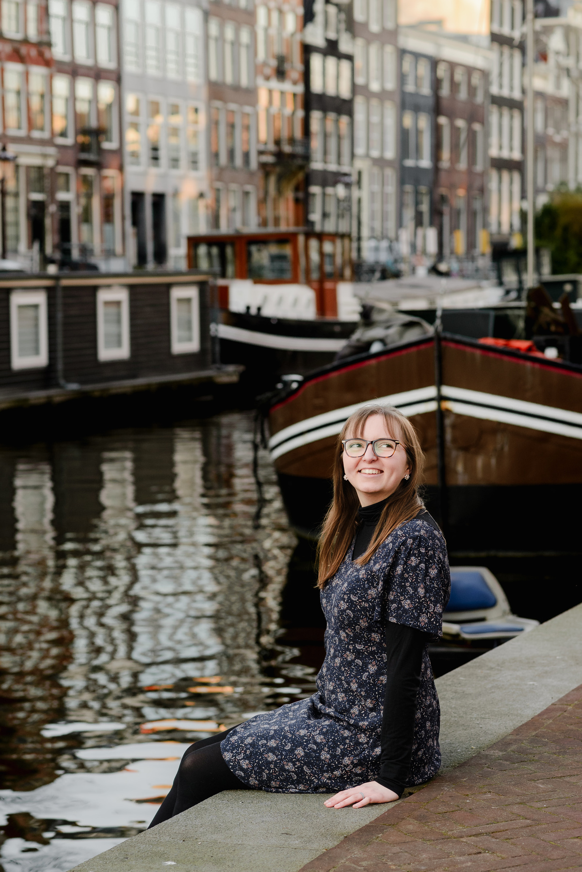 Individual portrait of the woman sitting beside an Amsterdam canal, with charming houseboats and reflections in the background.