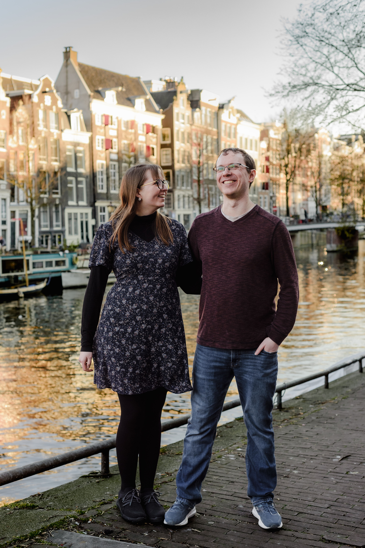 Smiling couple standing beside the canal in Amsterdam, surrounded by glowing reflections and classic Dutch townhouses—ideal for a casual anniversary photoshoot.