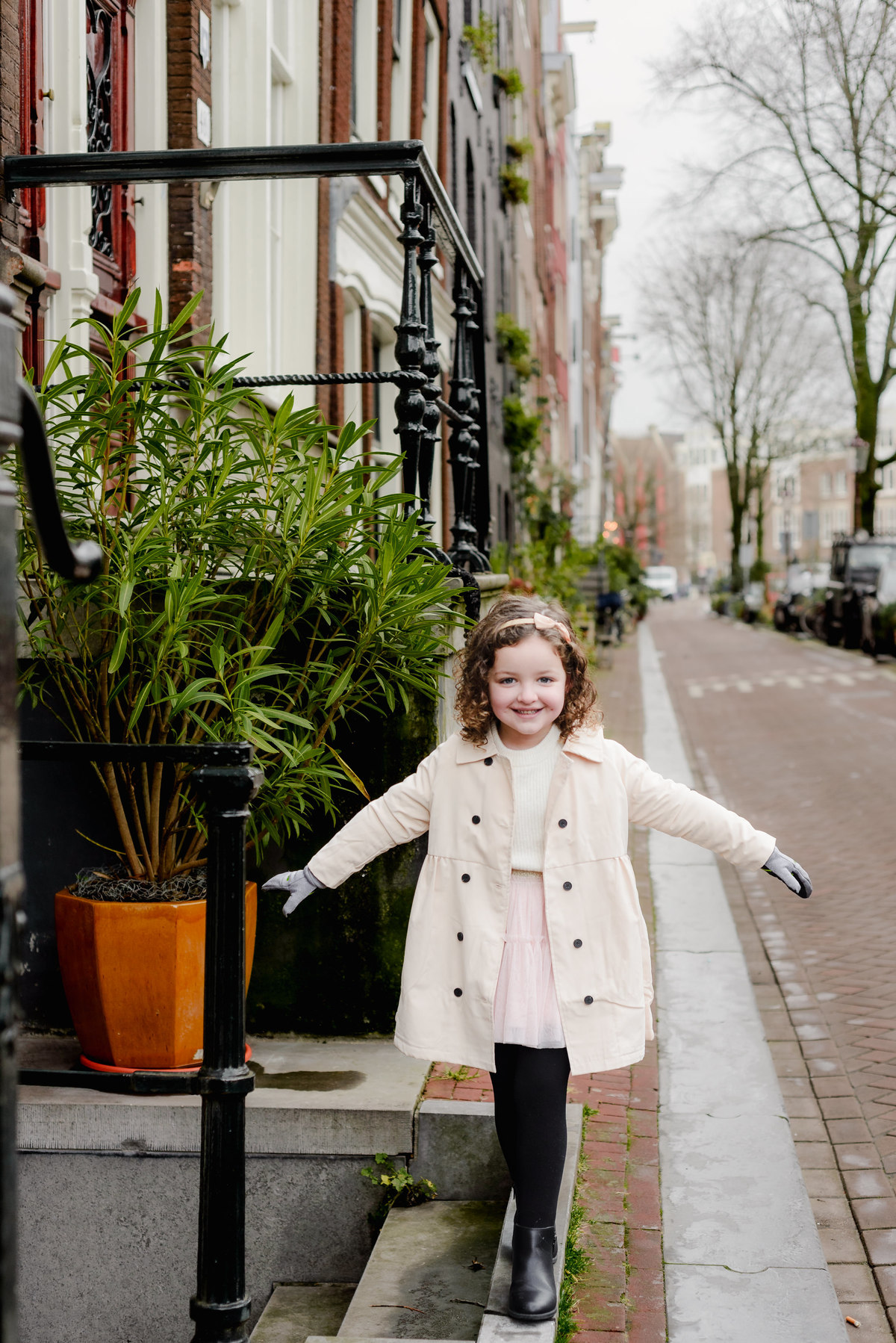 Giggling girl balances with outstretched arms along a classic Amsterdam street curb, surrounded by picturesque row houses.