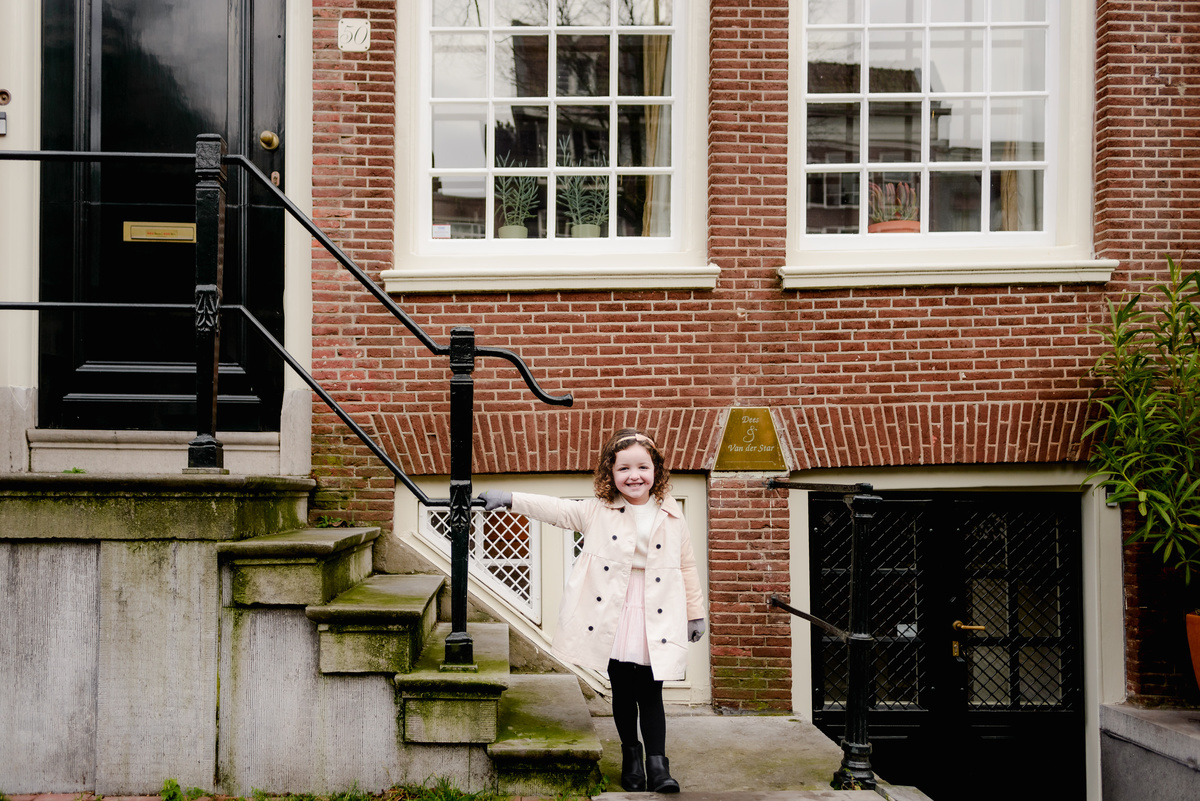 Smiling girl poses proudly on a historic Amsterdam doorstep, framed by red bricks and elegant iron railings.