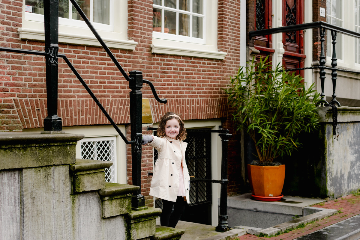 Young girl beams as she stands beside a classic Dutch staircase, the red-brick architecture of Amsterdam behind her.