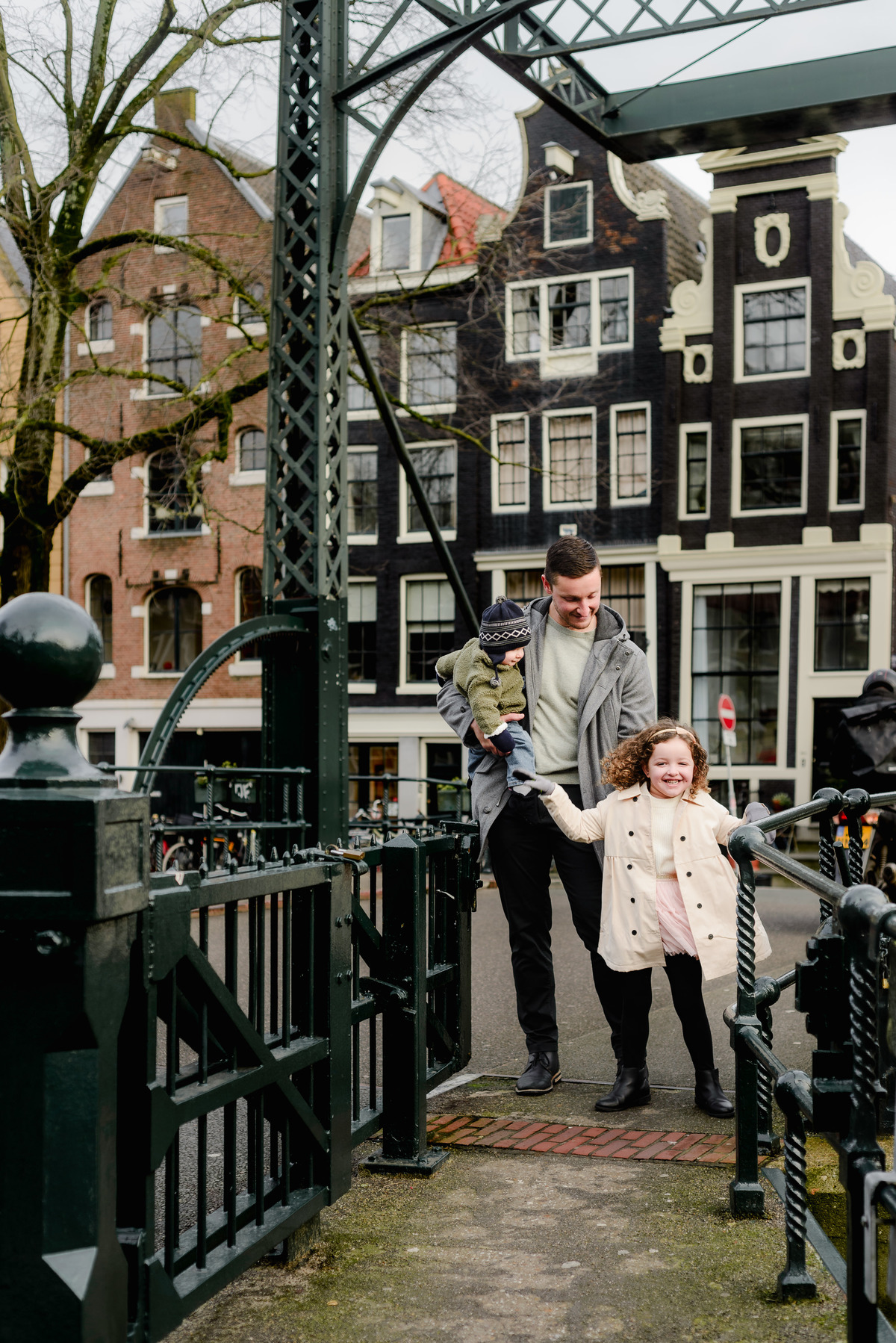 Dad holding baby and walking hand in hand with daughter across a classic Amsterdam iron bridge surrounded by historic homes.