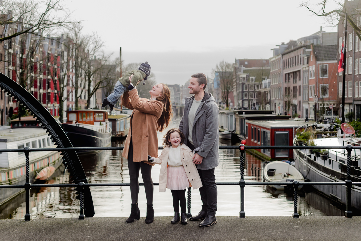 Mom laughs while holding baby and their daughter beams in front—capturing love and laughter along the Brouwersgracht bridge.