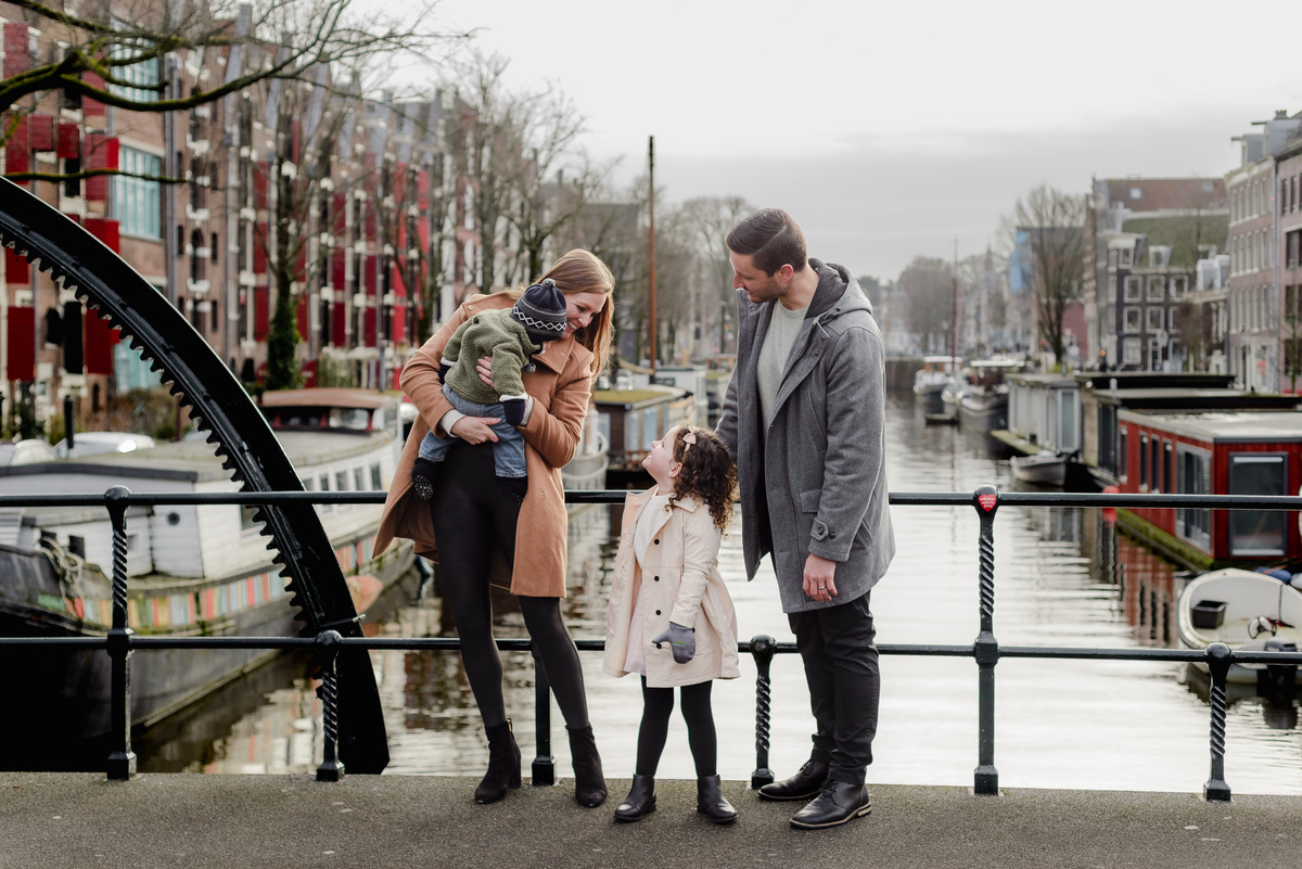 Parents share a kiss while their daughter twirls playfully in front, surrounded by colorful Dutch canal homes in the background.