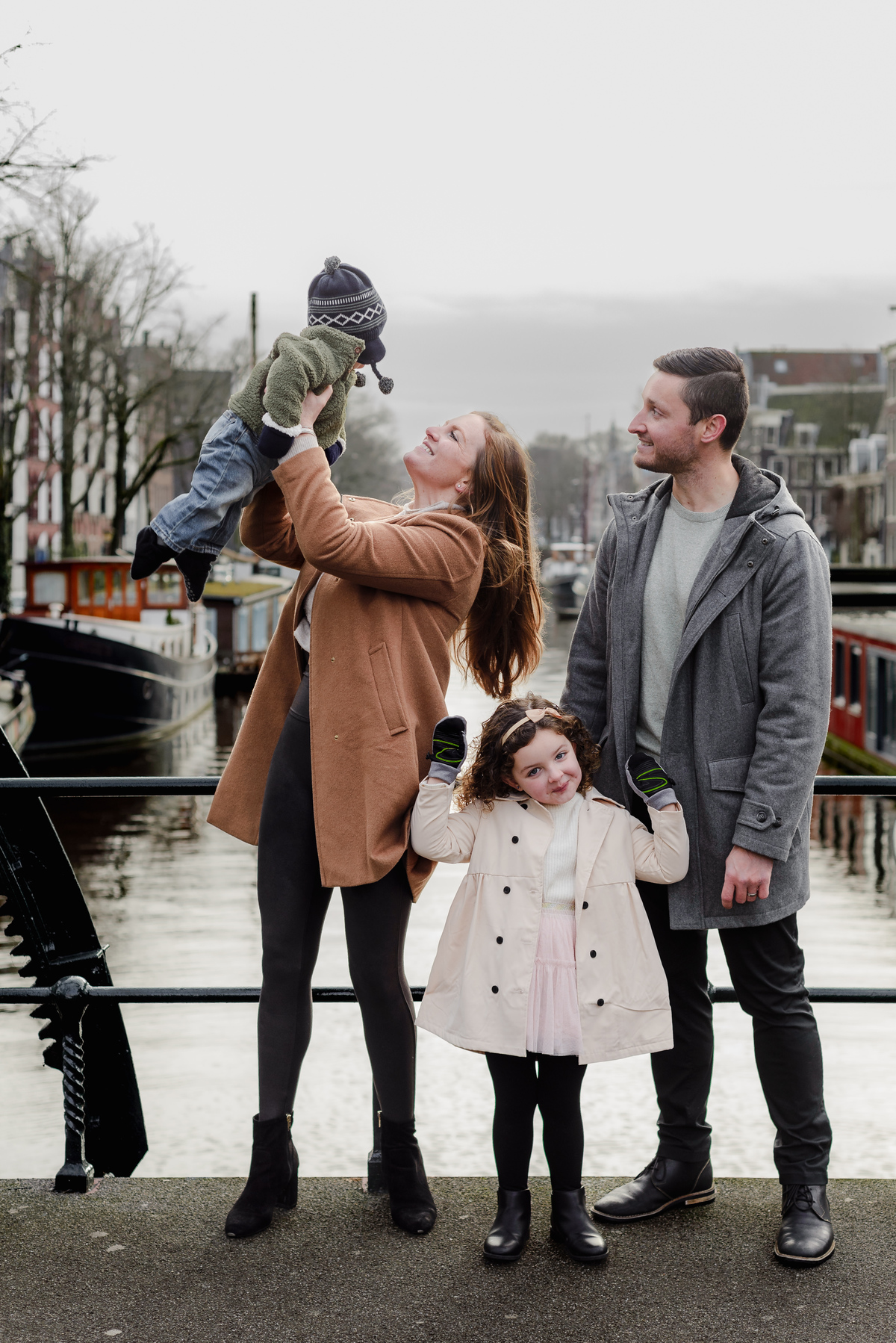 Mom and baby share a joyful moment as the family stands along a canal in Amsterdam, framed by iconic architecture and boats.