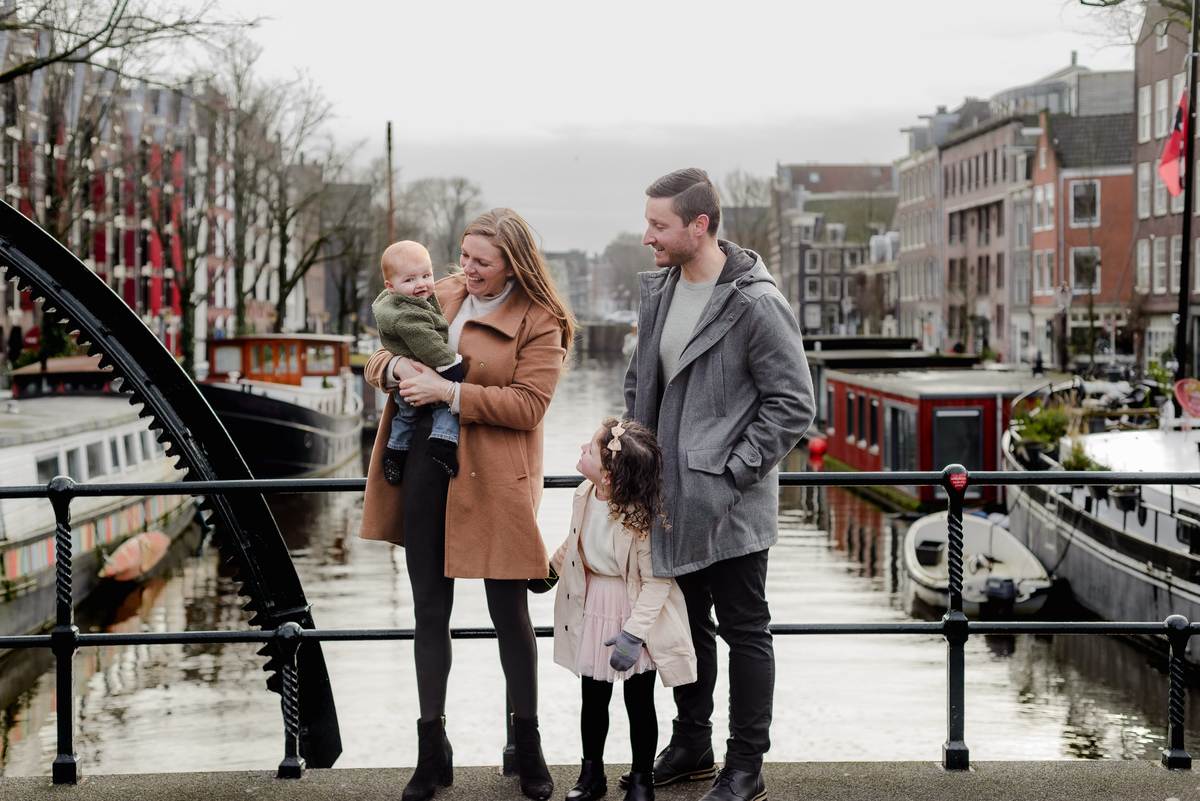 Smiling family of four on a charming canal bridge in Jordaan, Amsterdam, with houseboats and historic buildings behind them.