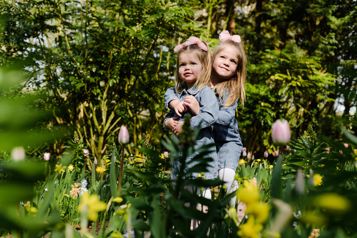 Two young sisters embrace among blooming tulips and daffodils in Keukenhof Gardens during early April, surrounded by lush spring greenery and vibrant Dutch flowers.