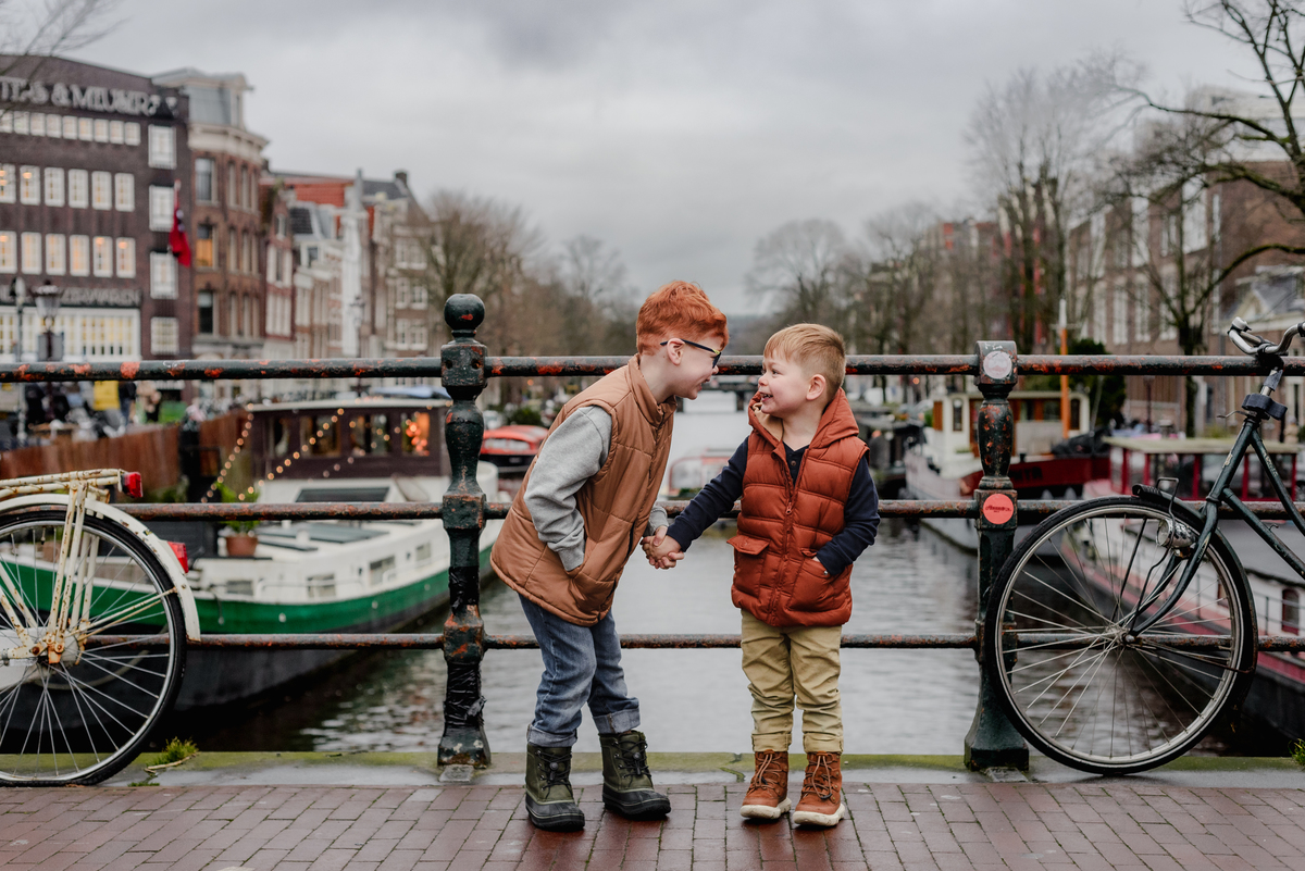 Two young brothers share a laugh while holding hands on a bridge in Amsterdam during a winter family photoshoot—set against a backdrop of canal boats, bikes, and historic Dutch architecture.