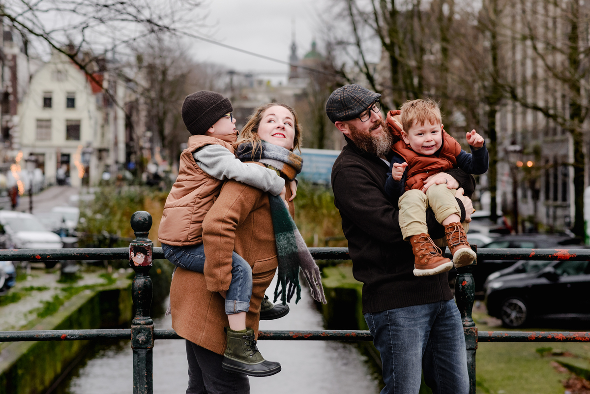 Parents carry their two young sons during a December family photoshoot in Amsterdam, set against a peaceful canal and narrow street backdrop, capturing candid connection and winter charm in the city center.

