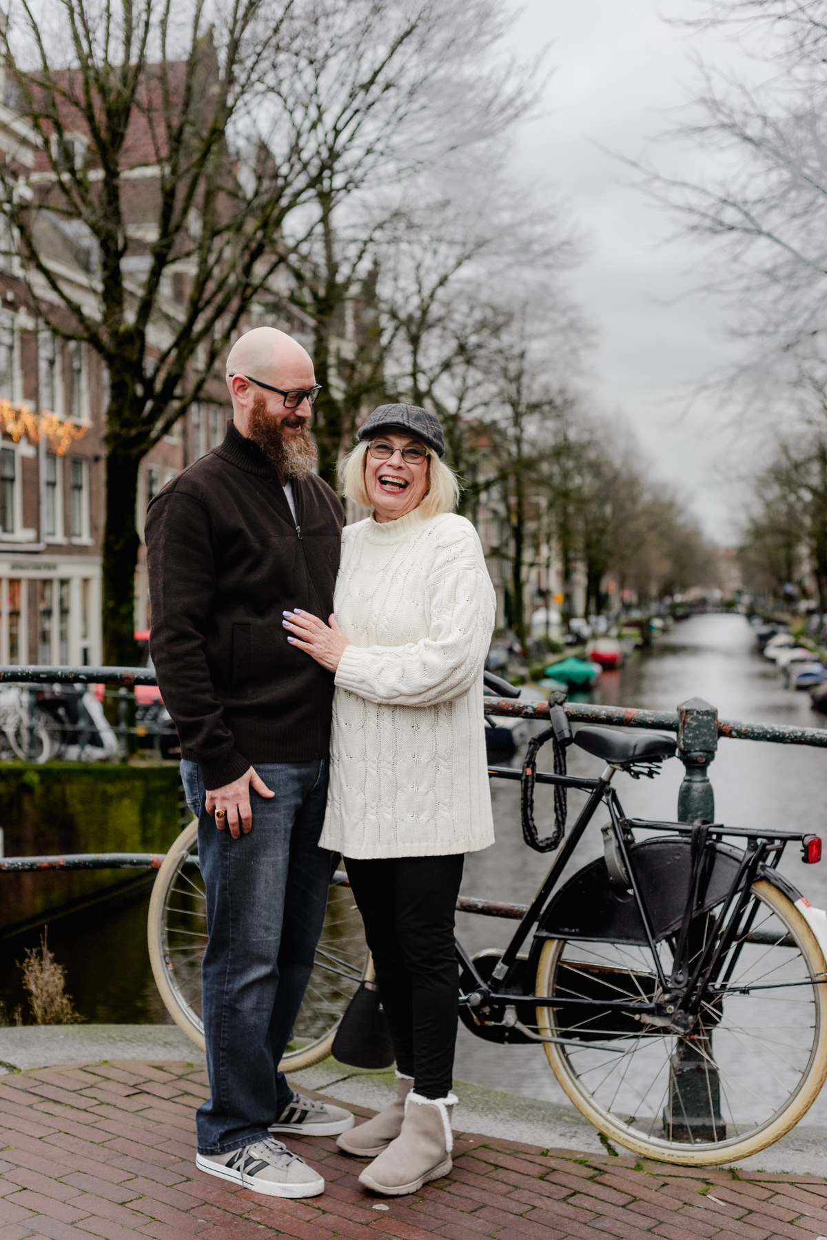 A mother laughing next to her adult son on an Amsterdam canal bridge during a December photo session, surrounded by bicycles and historic canal houses—capturing a tender moment in the heart of the city.
