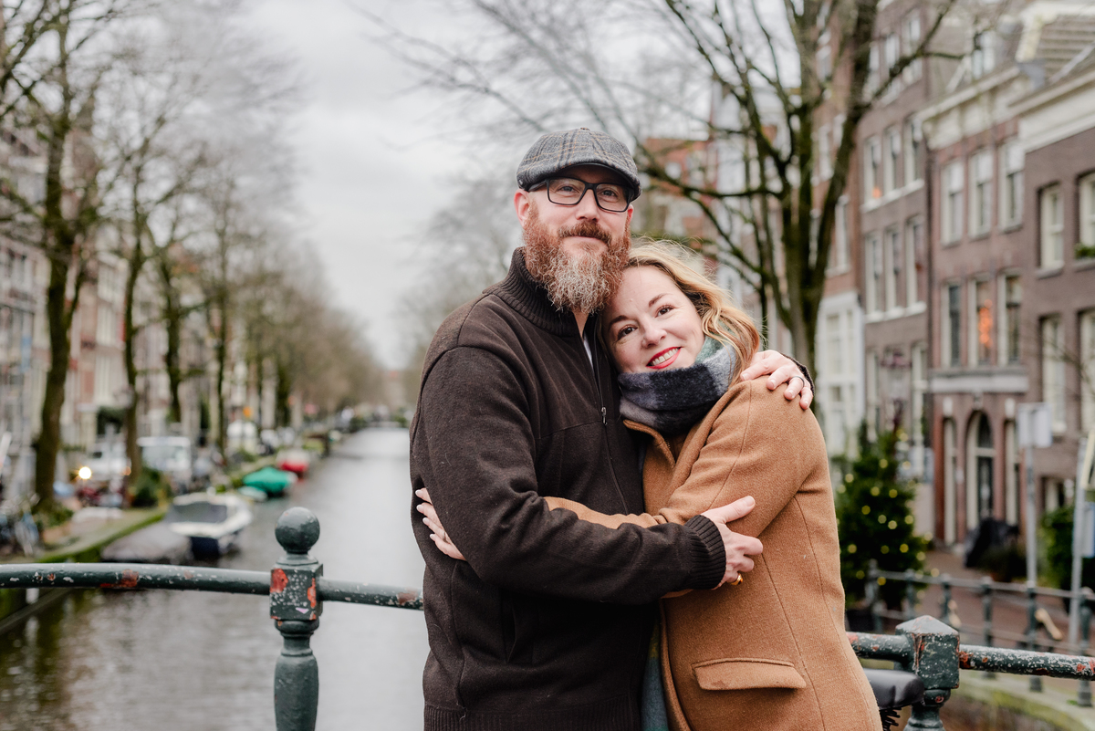 A couple embraces on an Amsterdam canal bridge during a cozy December photo session, with historic buildings, moored boats, and soft winter light creating a romantic city backdrop.