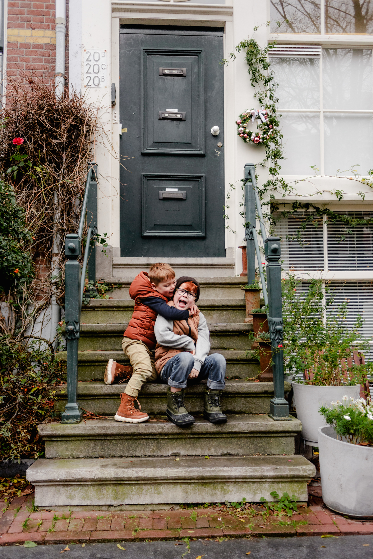 Two brothers sit on the steps of a traditional Amsterdam home during a December family photoshoot—surrounded by ivy, winter greenery, and a festive wreath, capturing the charm of Dutch architecture and candid sibling connection.