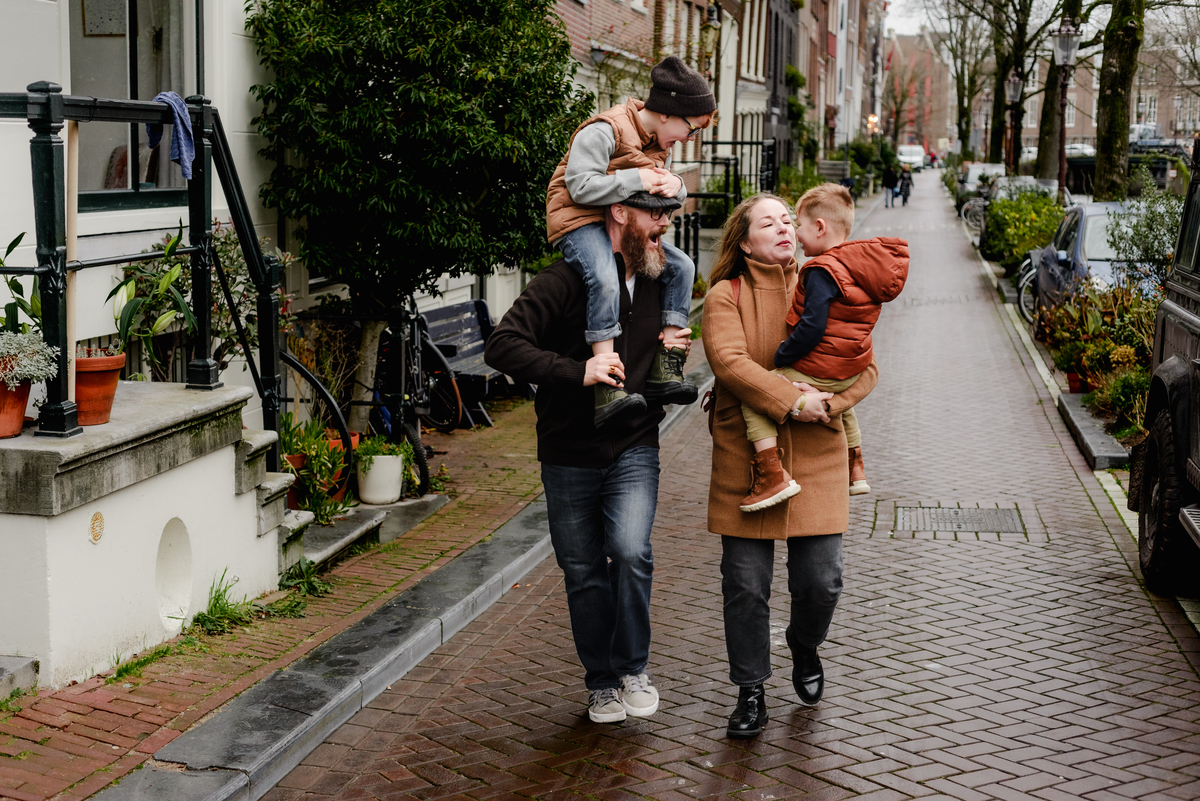 A family walks together through a charming Amsterdam street during a December photo session—dad carrying one child on his shoulders and mom holding the other, capturing natural joy and city charm in a candid lifestyle moment.