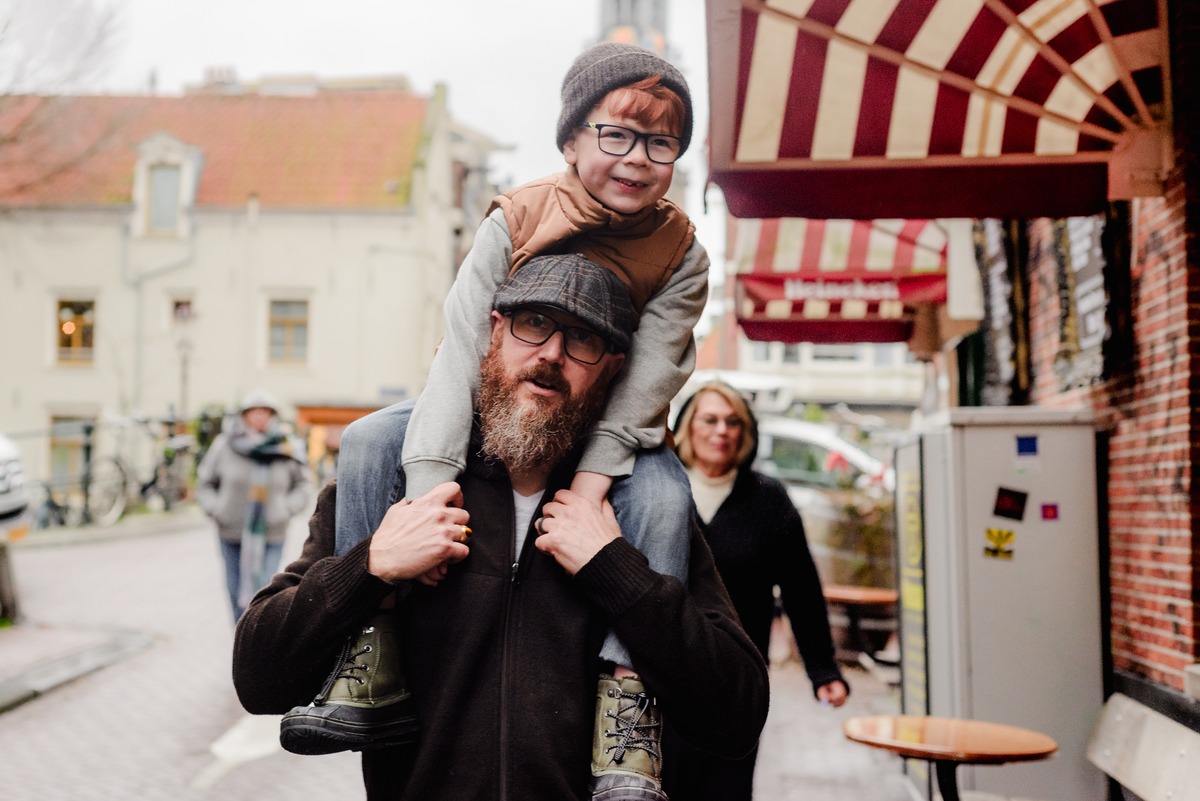 A father carries his son on his shoulders through a cozy Amsterdam street during a winter family photo session, with striped awnings, brick buildings, and Dutch charm creating the perfect backdrop.