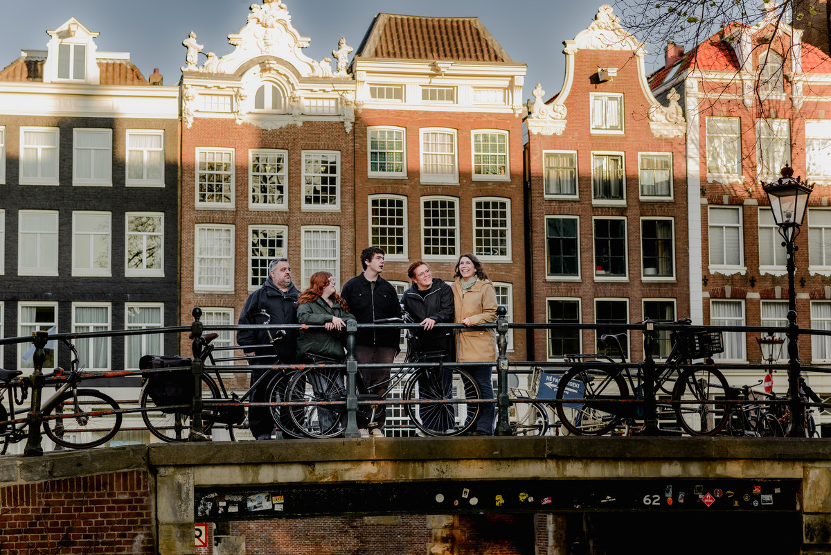 Family standing on a bridge over a canal, framed by bikes and historic Amsterdam townhouses, smiling and looking at one another.