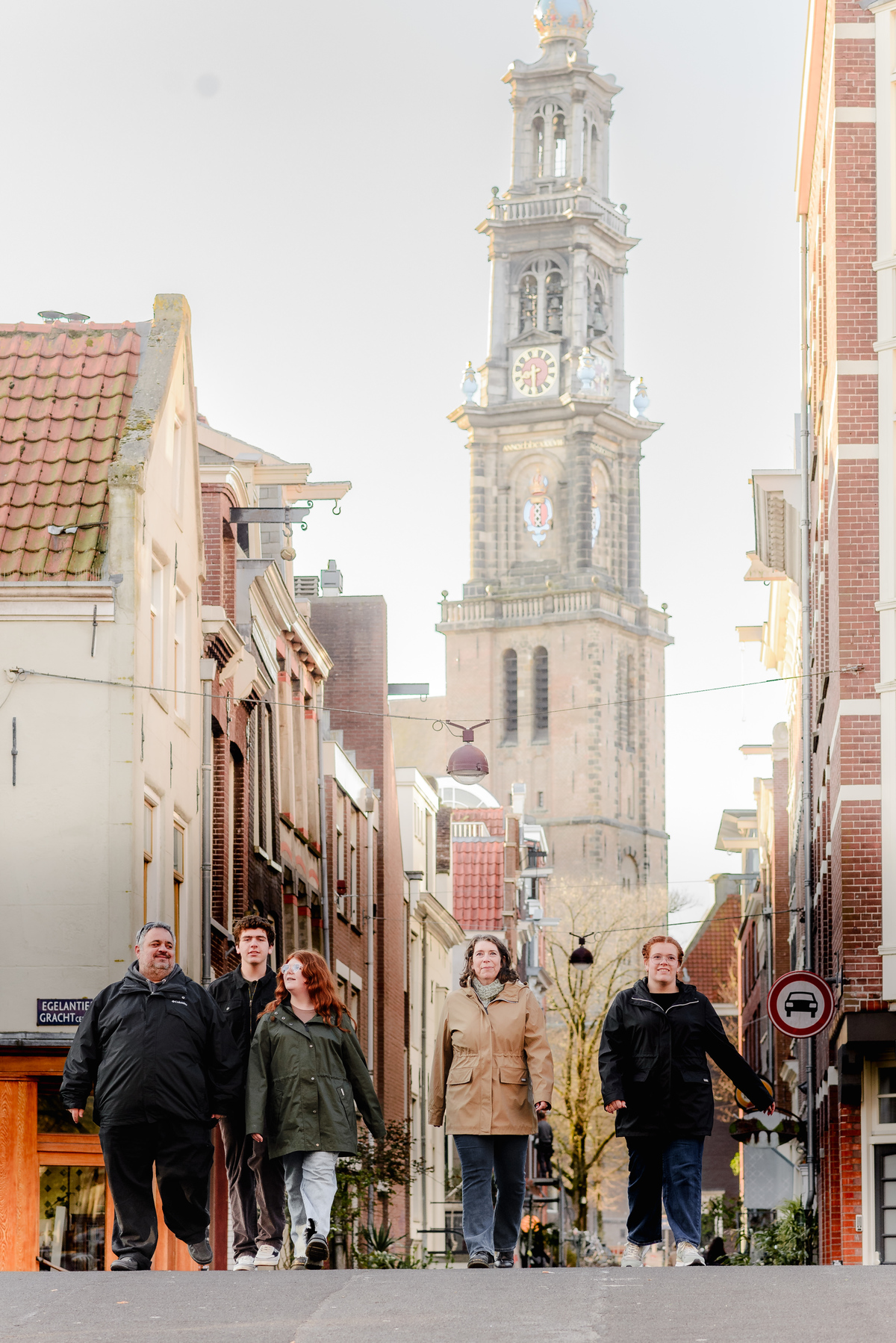 Family of five walking toward the camera on Egelantiersgracht street, with the iconic Westerkerk tower rising in the distance.