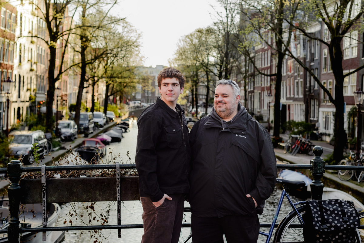 Father and teenage son standing side by side on a canal bridge, bathed in warm morning light with houseboats and trees in the background.