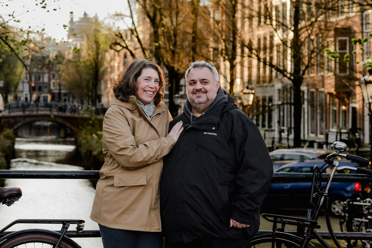 Couple smiling warmly while standing on a bridge with a canal behind them, surrounded by spring trees and golden light.