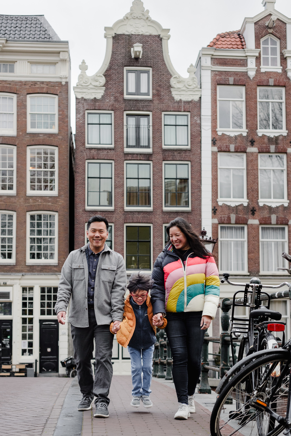 Parents walking hand-in-hand with their son on a canal bridge, playfully lifting him in the air during a joyful family moment.