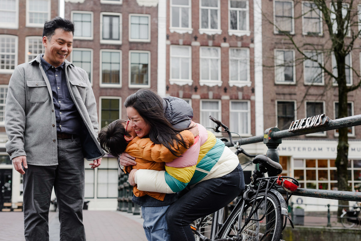 Mother hugging her son tightly with a big smile, while father looks on with joy — captured in front of iconic Amsterdam canal houses.