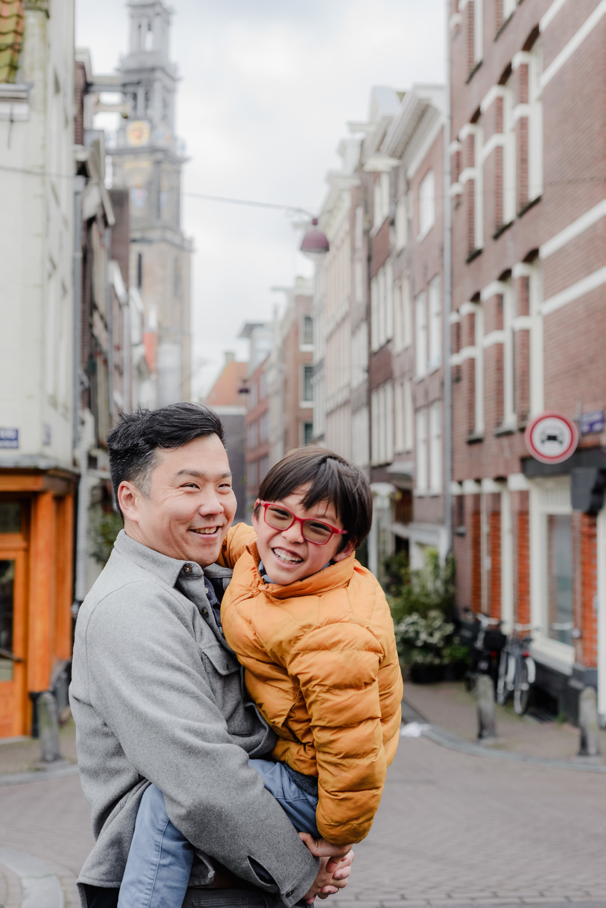Father holding his son with both smiling brightly, set on a quiet Amsterdam street with Westerkerk in the distance.