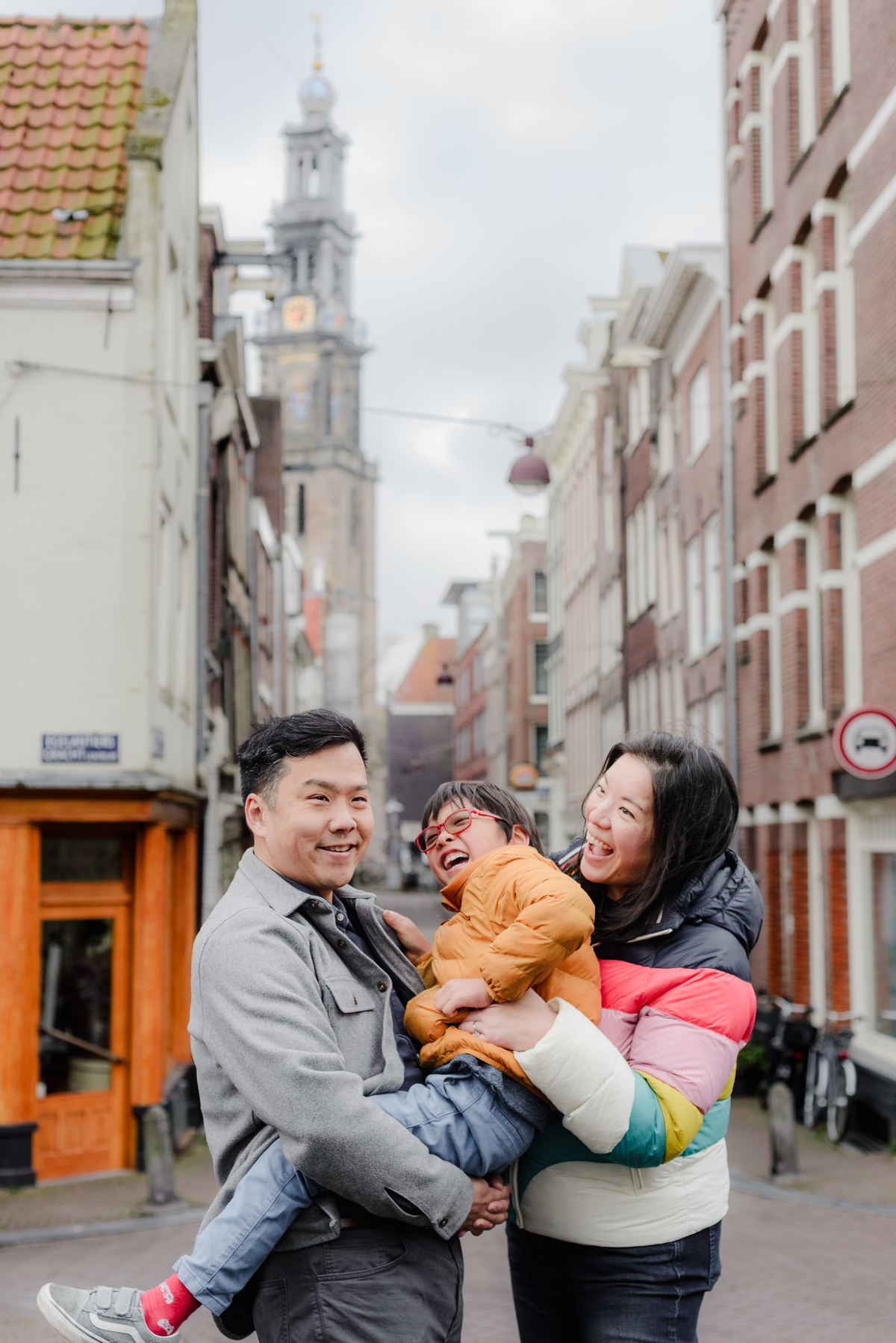 Playful family moment with both parents holding their son, laughing together under cloudy skies on an Amsterdam street.