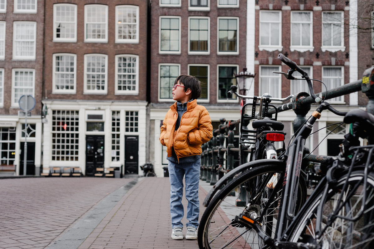 Young boy in an orange jacket standing on a canal bridge in Amsterdam, looking to the side with classic Dutch buildings behind him.