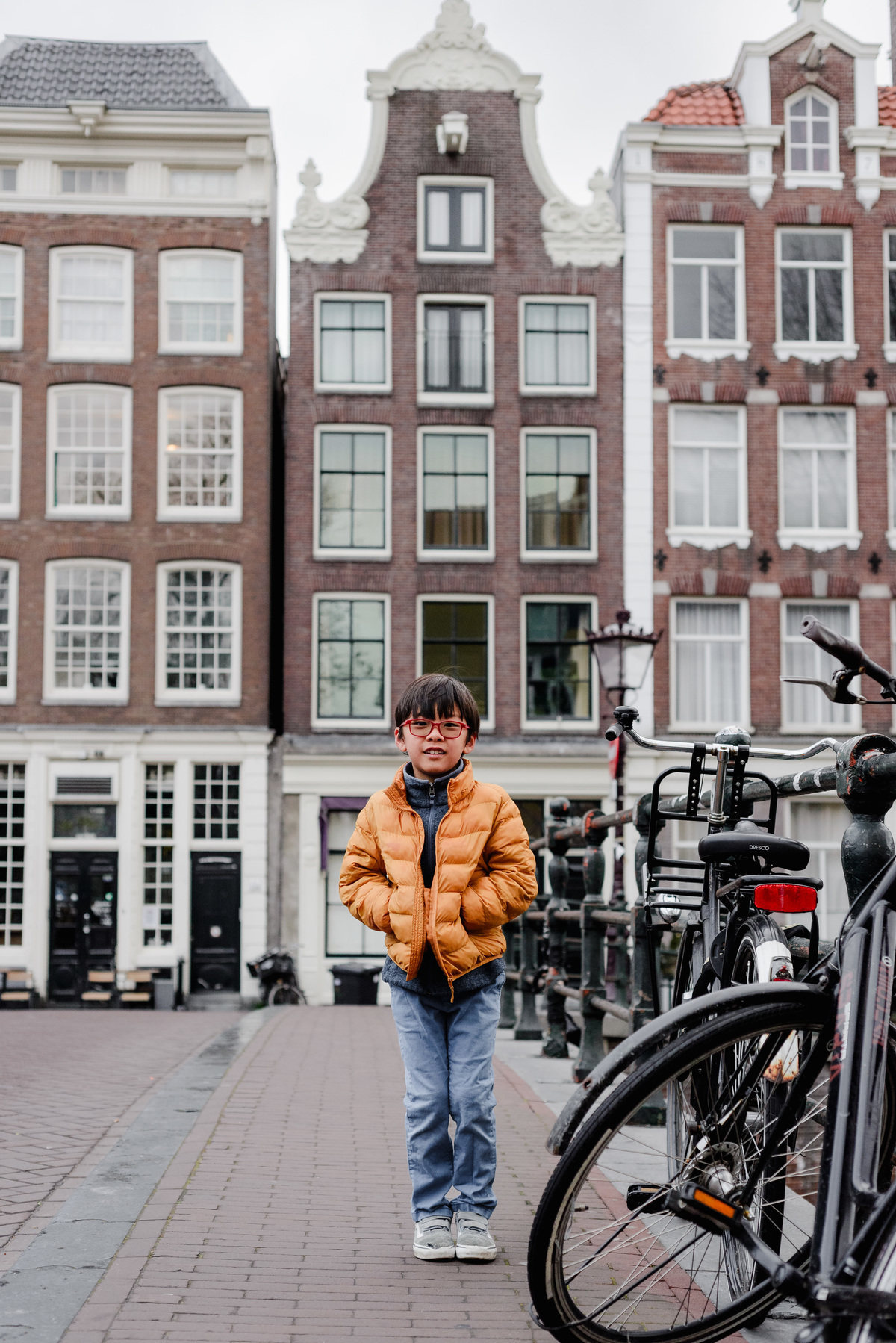 Portrait of a smiling young boy in an orange puffer jacket standing confidently on an Amsterdam bridge, framed by bicycles and tall gabled houses.