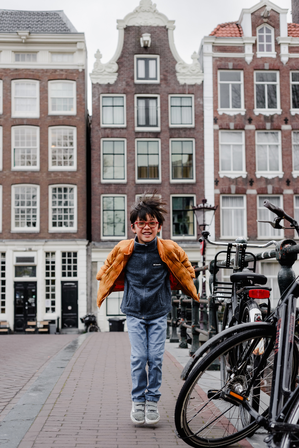 Boy mid-jump with wind-blown hair, smiling joyfully on a quiet Amsterdam street lined with traditional canal houses.