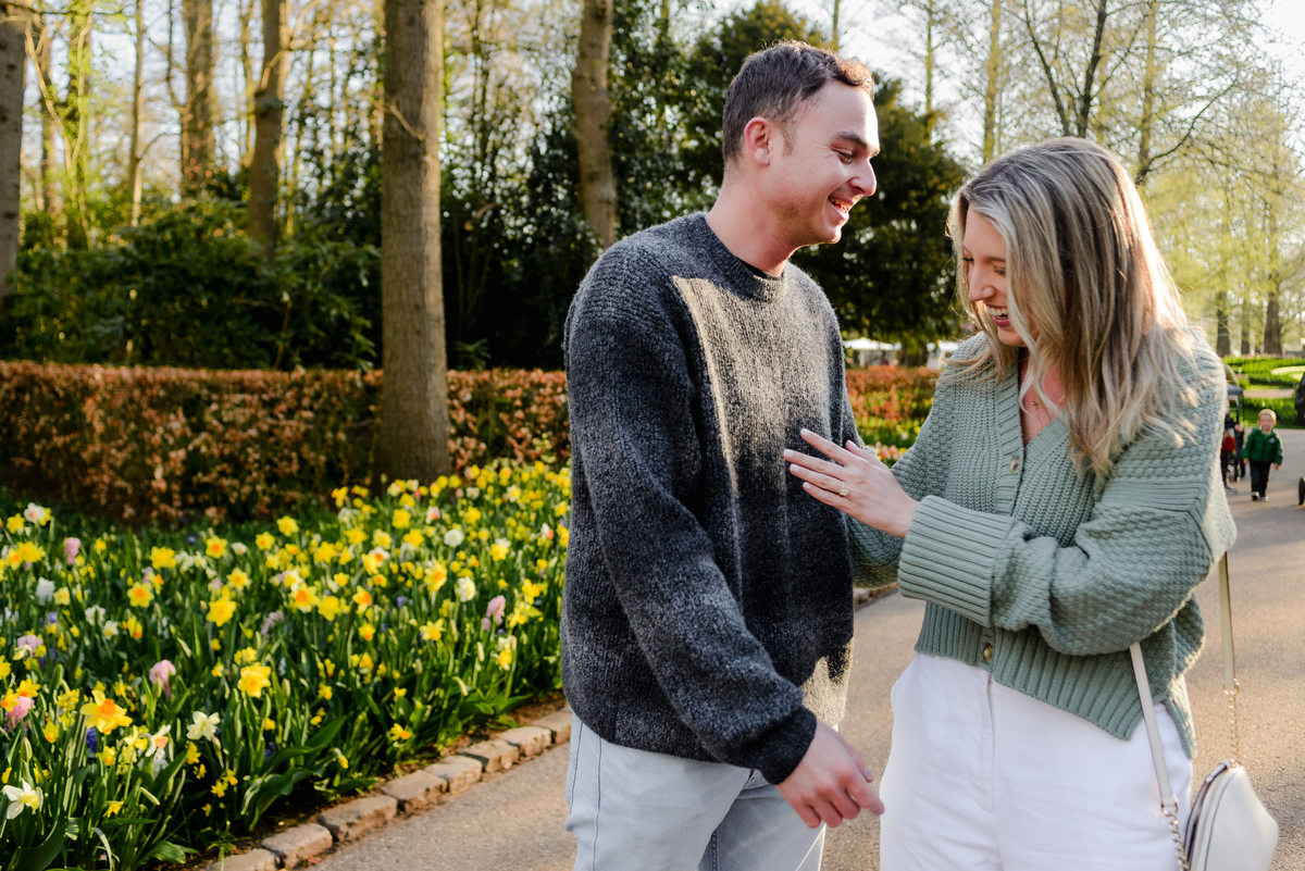 Smiling bride-to-be shows off her engagement ring, standing hand-in-hand with her fiancé in a garden walkway.
