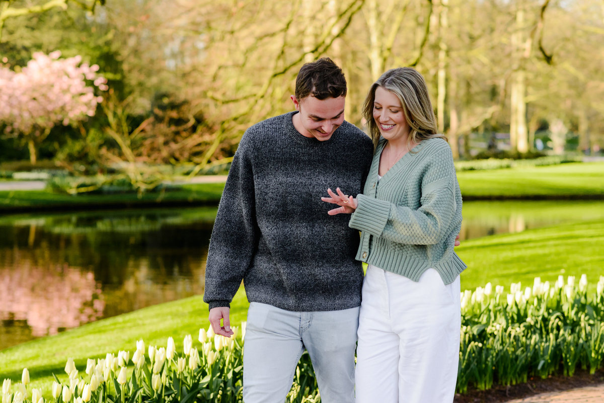 Engaged couple walking beside a serene pond, smiling as she admires her new ring under the morning sun.