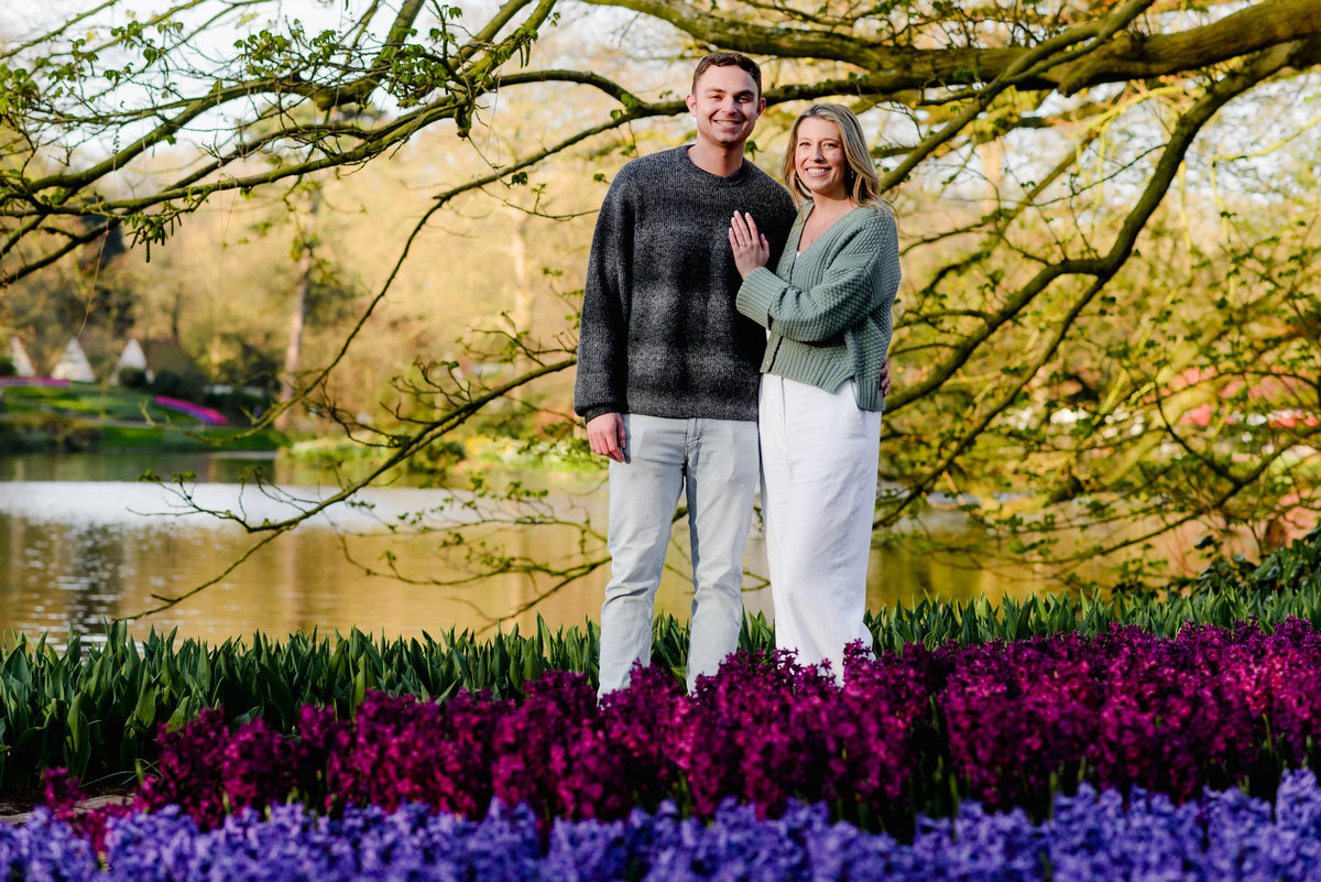 Posed portrait of the newly engaged couple standing together in front of a lake and vibrant purple hyacinths.