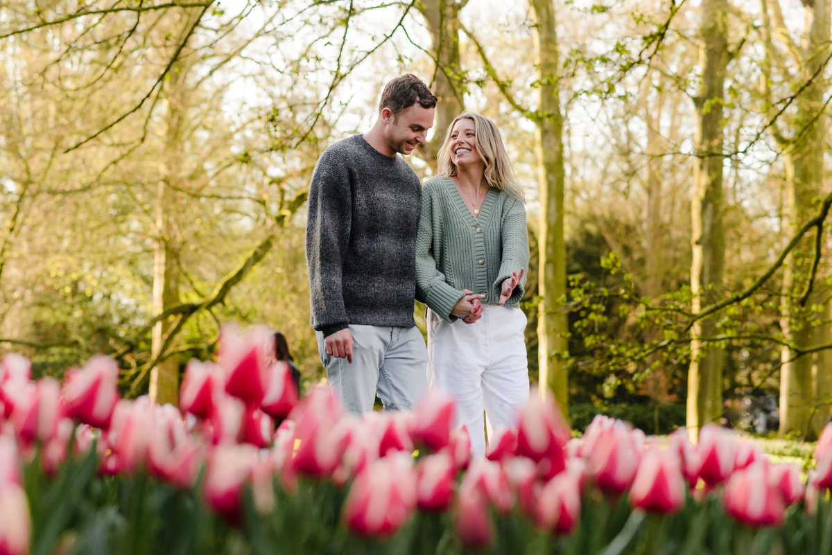 Couple walking hand-in-hand past a vibrant field of tulips, exchanging loving glances after their engagement.