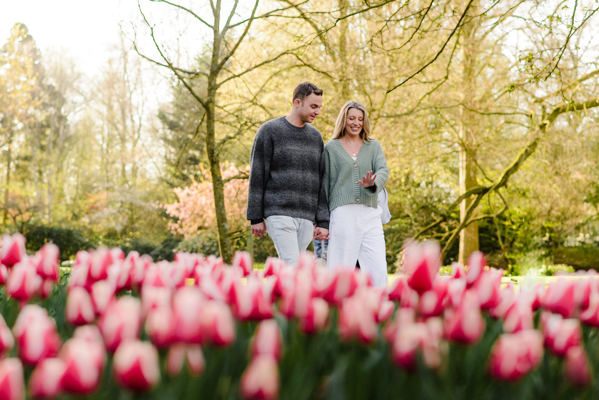 Bride-to-be looking at her engagement ring as the couple walks through tulip-lined paths, smiling with joy.