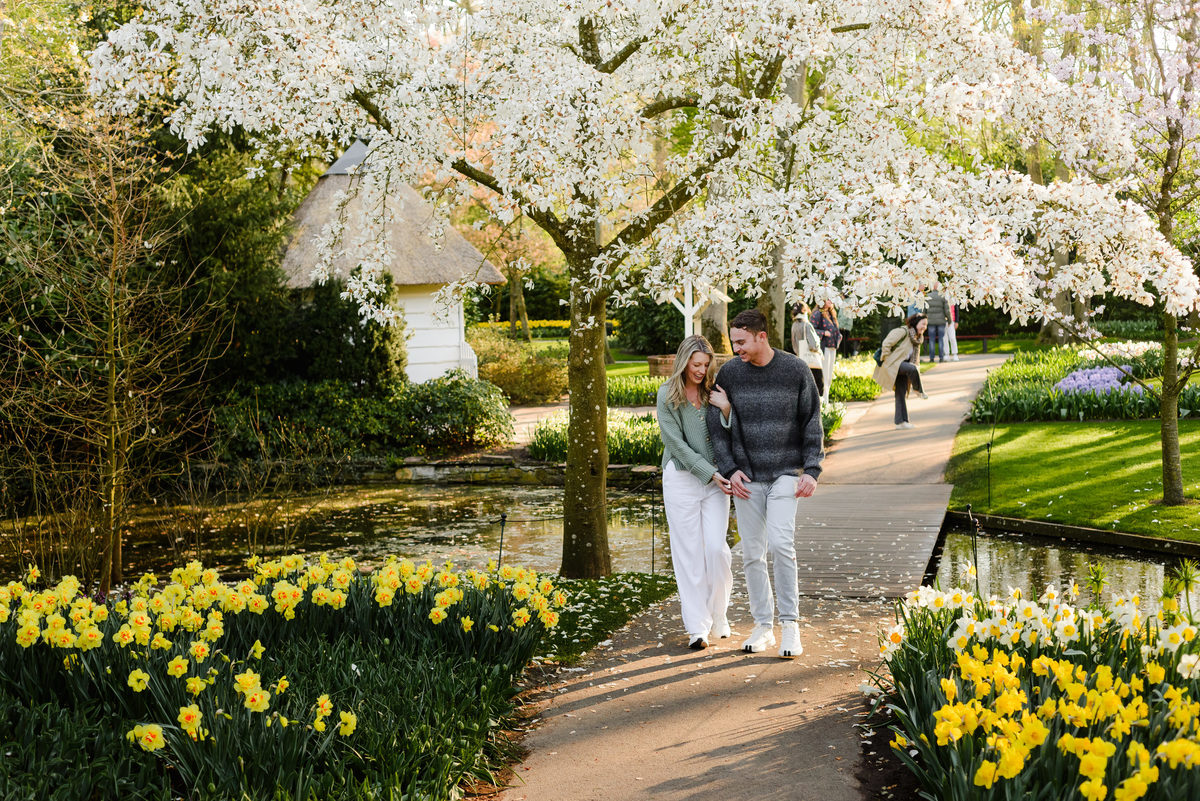 Romantic stroll beneath a blooming magnolia tree, with yellow daffodils and a quaint garden cottage nearby.