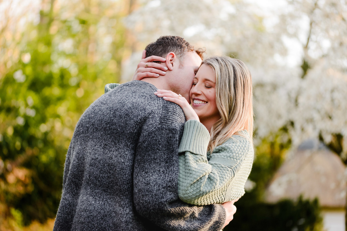 Couple embraces tenderly, her engagement ring visible as she smiles with joy under blooming spring trees.