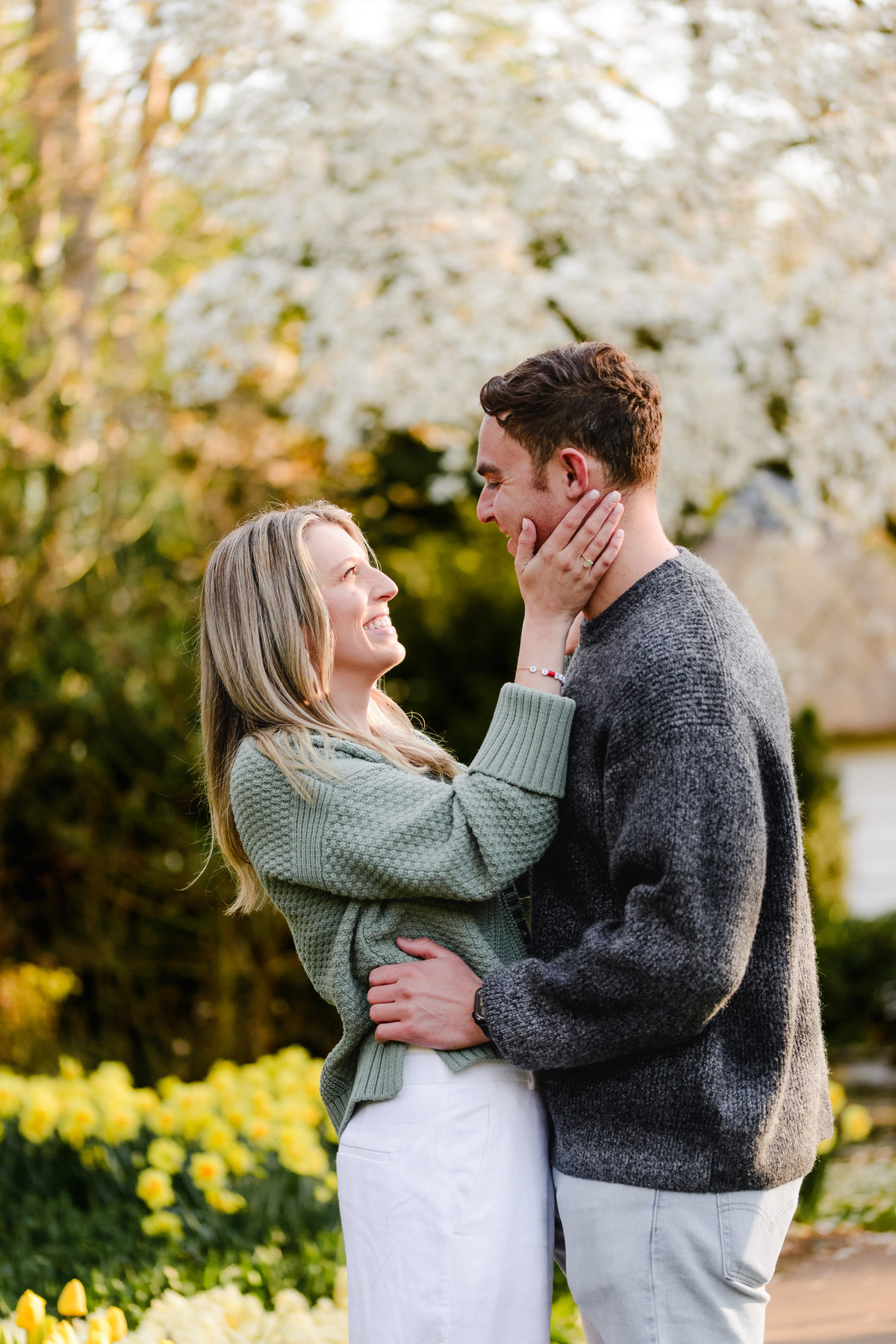 Engaged couple gazes lovingly at each other with blooming white trees and yellow tulips in the background.