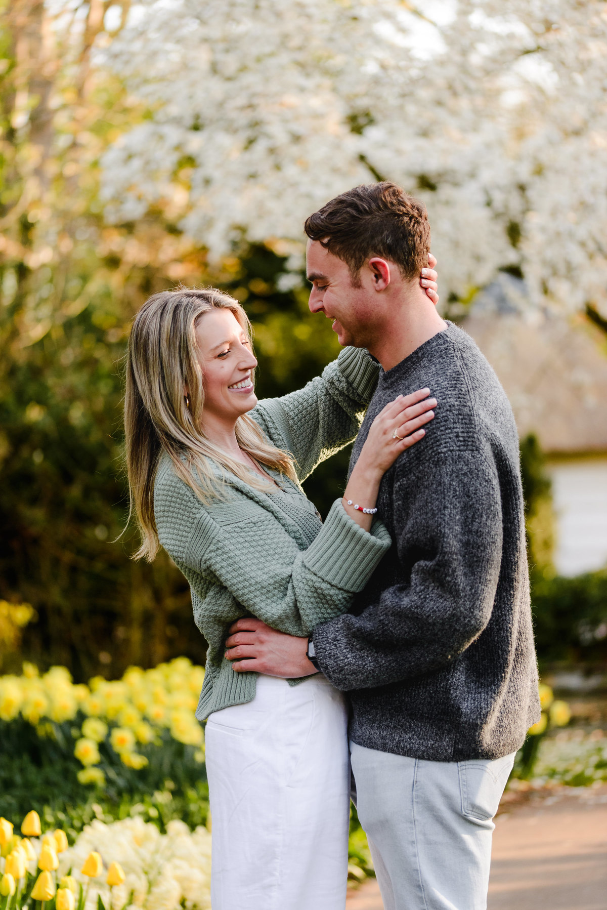 Fiancée gently touches her partner’s face as they laugh together in a garden full of spring flowers.