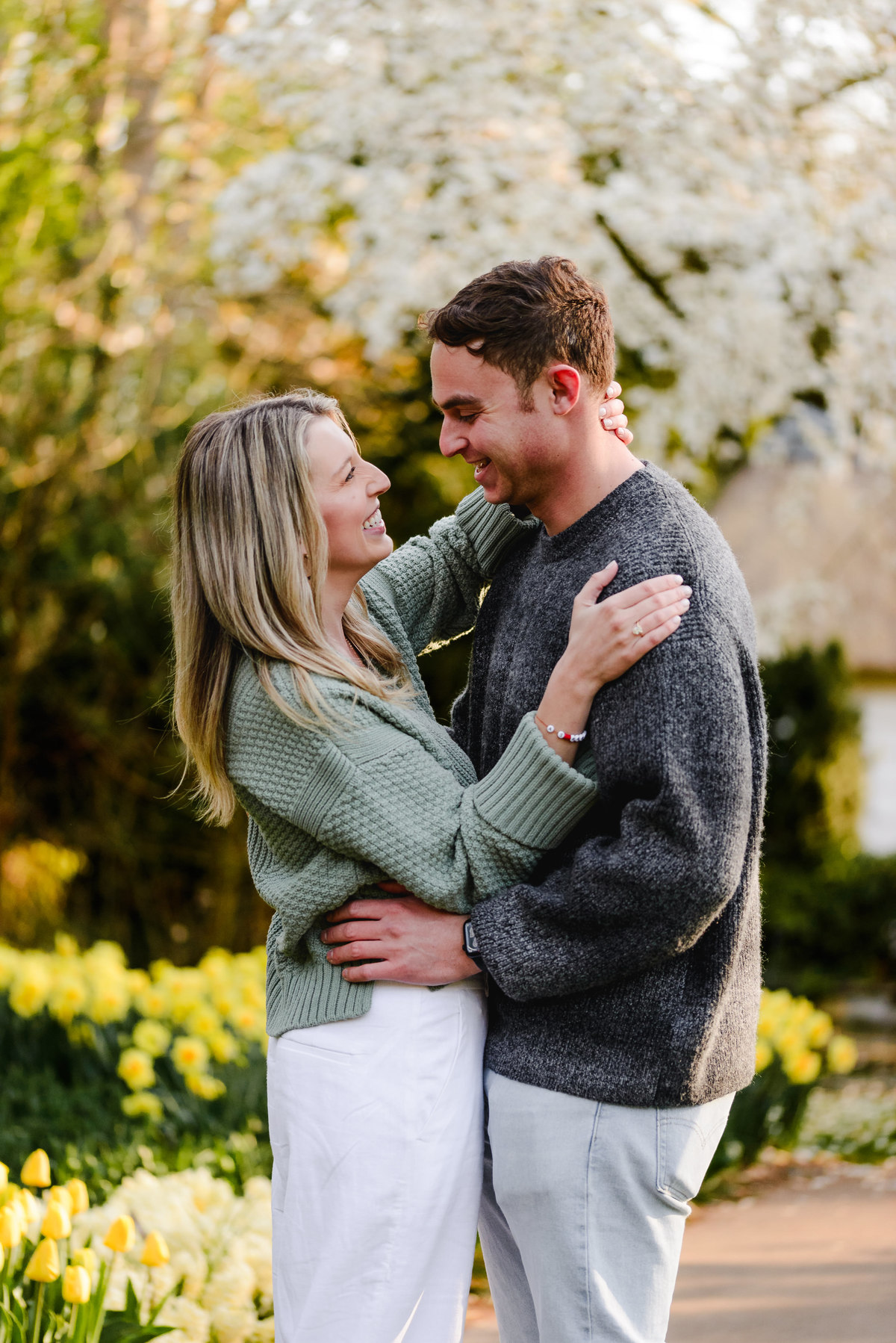 Romantic moment between the newly engaged couple, smiling under flowering trees in a lush park setting.